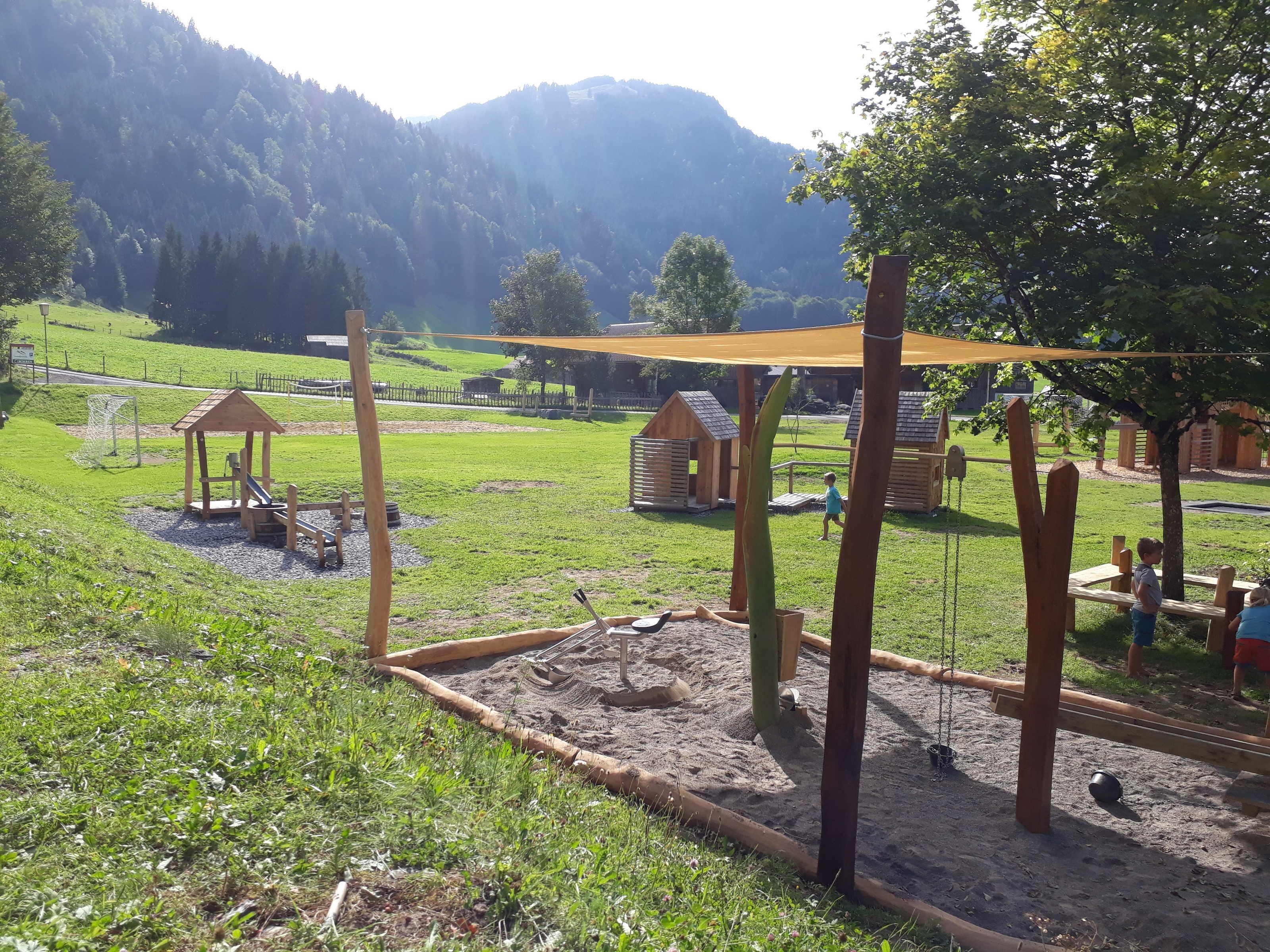 Un parque infantil con estructuras de madera, zona de juegos con arena y un campo de césped con montañas al fondo.