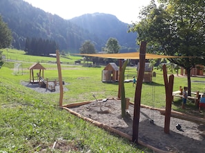 A playground with wooden structures, a sandy play area, and a grassy field with a mountain backdrop.