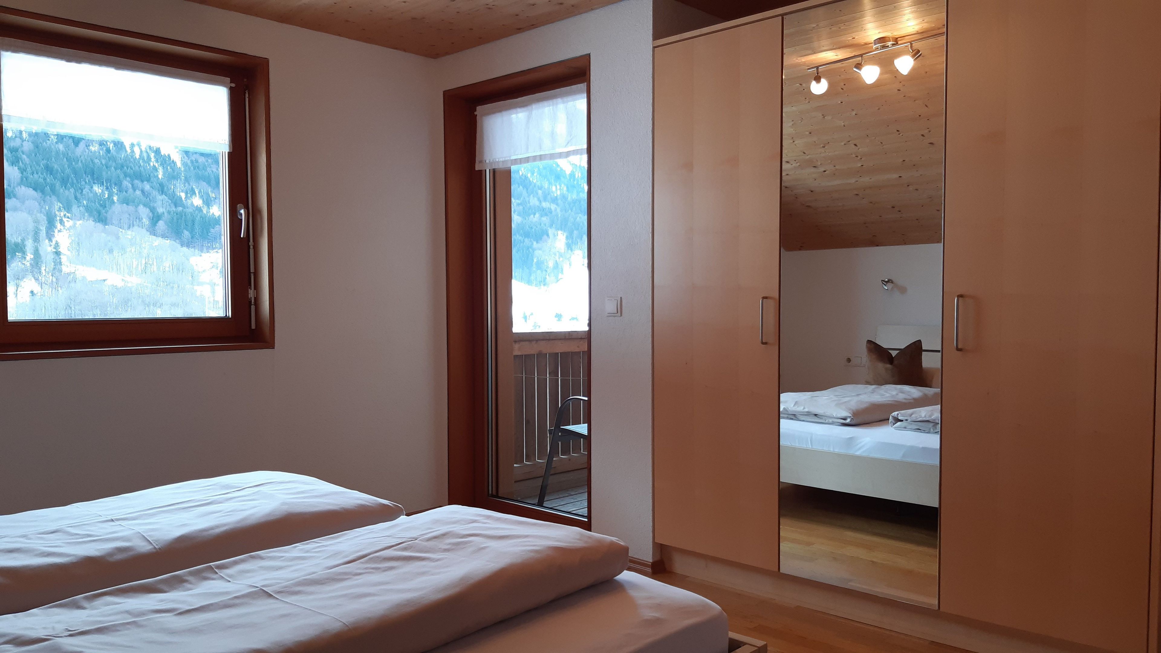 A bedroom with two beds, a wooden wardrobe, a window with a view of a snowy landscape, and a balcony with a chair.