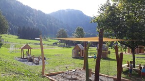 A playground with wooden structures, a sandy play area, and a grassy field with a mountain backdrop.