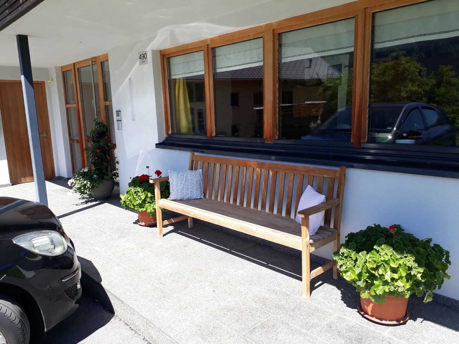 A wooden bench outside a building with large windows, a car parked nearby, and potted plants.