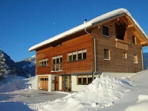Un chalet de madera de dos pisos con un techo cubierto de nieve, con montañas al fondo.