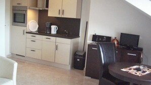 A compact kitchen with a microwave, stove, and coffee maker. There is a dining table with chairs and a television in the corner. - VV 32; 2-space; 1ST FLOOR; Balcony; 38m² - Villa Vogelsang (Sierksdorf)