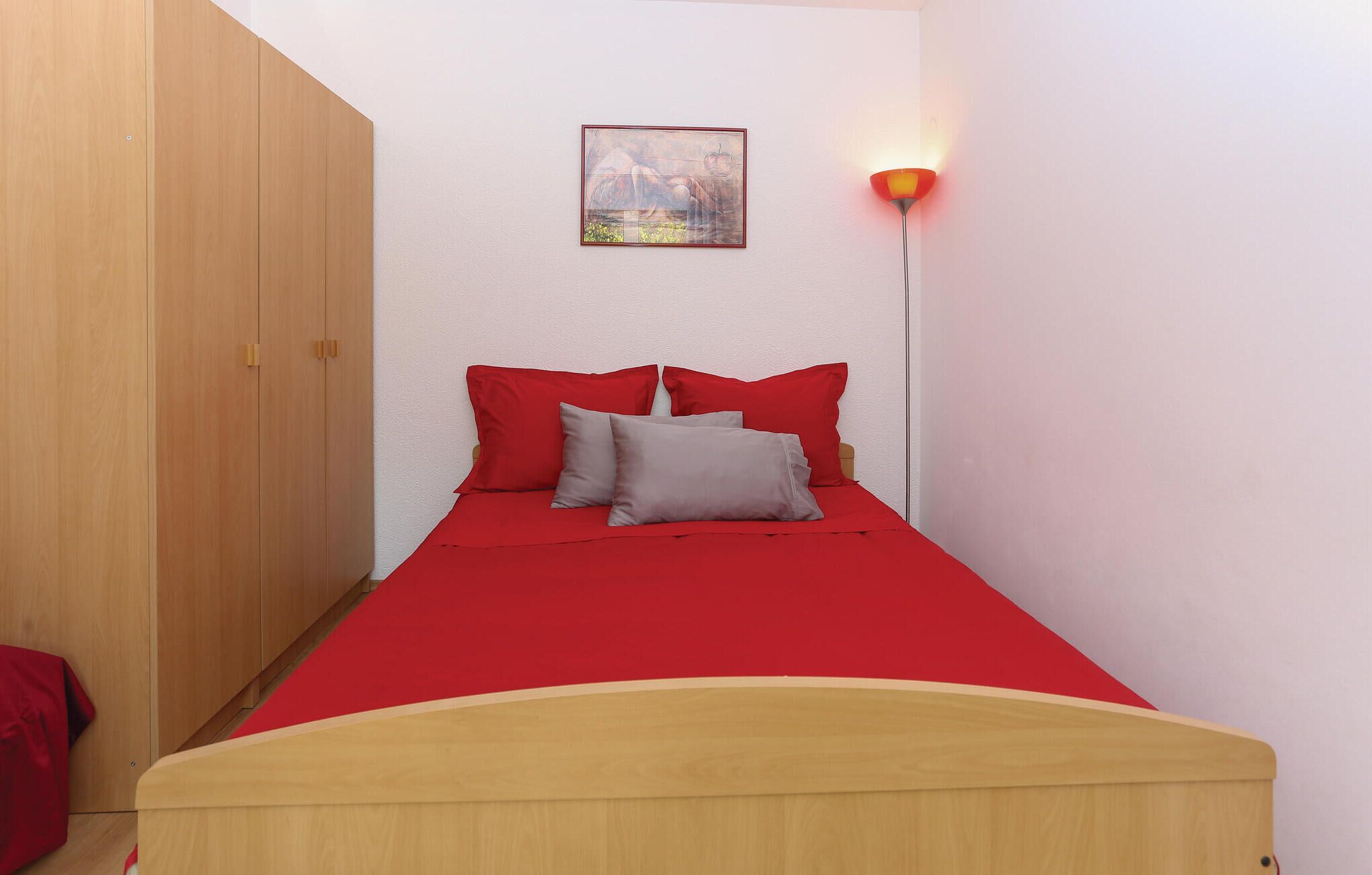 Una habitación pequeña con una cama, un armario de madera, un cuadro colgado en la pared y una lámpara de pared.