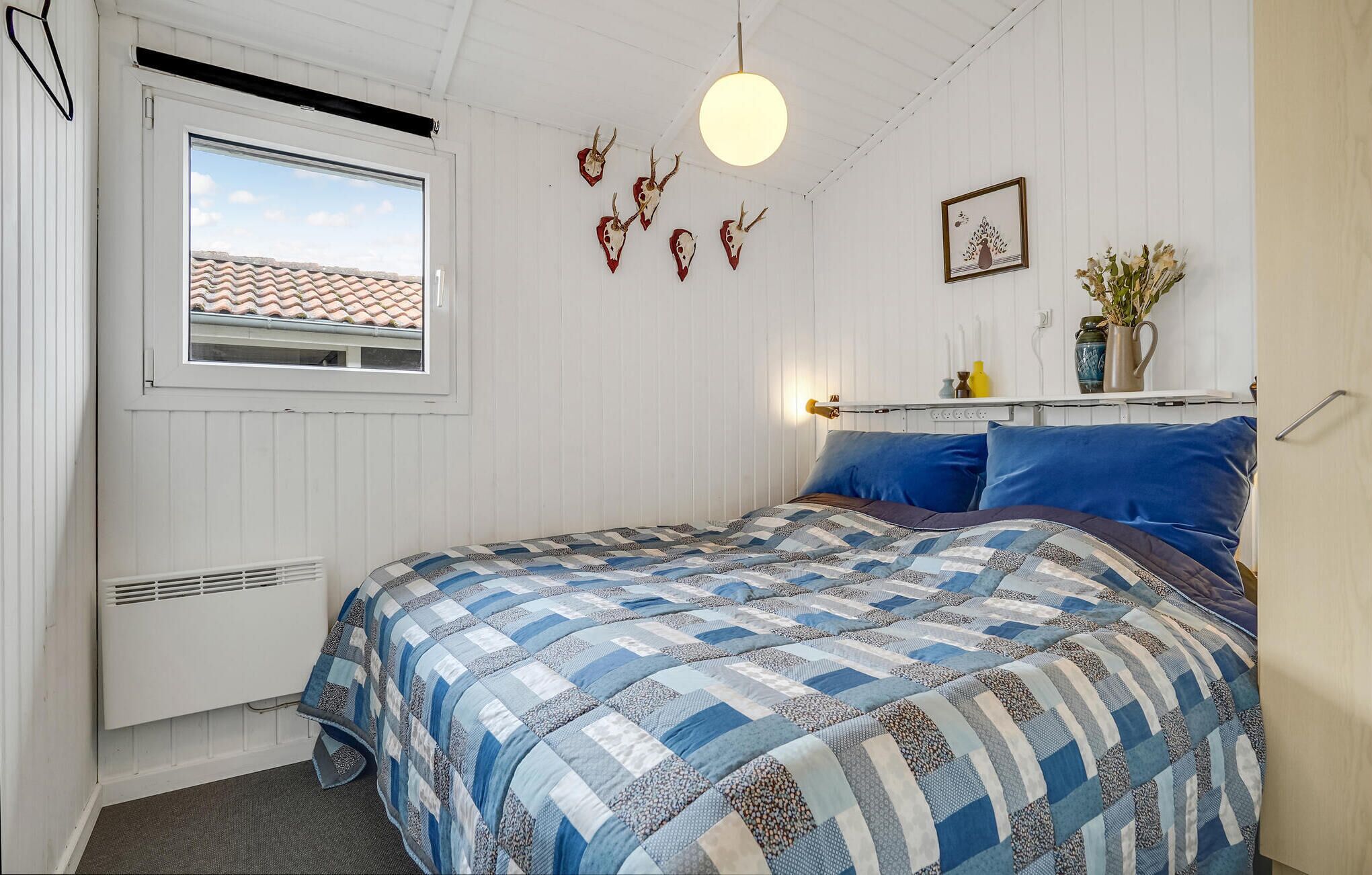 Quarto com cama, janela, radiador, prateleira na parede com objetos decorativos e luminária no teto.