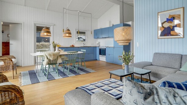 Un salón amplio con un sofá gris, una zona de comedor con una mesa y sillas blancas, y una cocina con armarios azules y una estufa blanca.