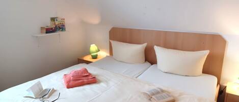 Un lit double avec du linge de lit blanc, deux oreillers, une lampe de chevet et une table de chevet avec une lampe et des livres.