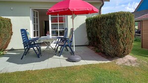 Een terras met een rode parasol, twee blauwe stoelen, een kleine tafel met een bloempot en een haag.
