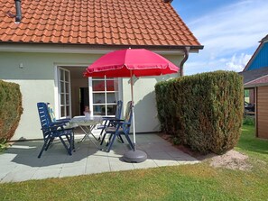 Une terrasse avec un parasol rouge, deux chaises bleues, une petite table sur laquelle est posée une plante en pot et une haie.