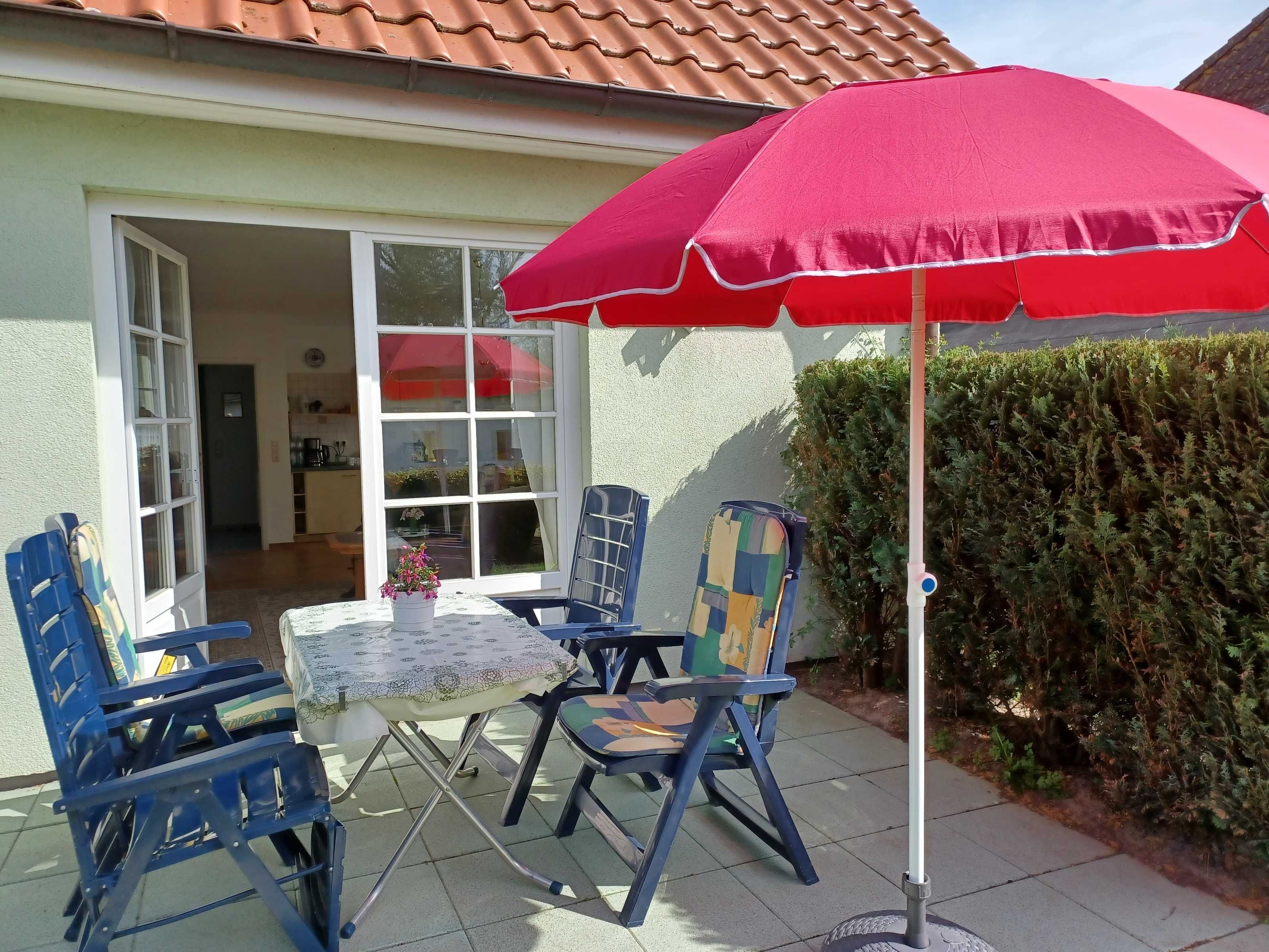 An outdoor patio with a table and chairs, a red umbrella, and a hedge.