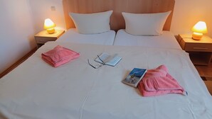 Een net opgemaakt bed met witte lakens, twee kussens, een paar brillen en een boek. Twee bedlampen staan op houten nachtkastjes.
