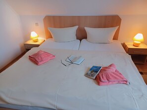 Un lit bien fait, avec du linge de lit blanc, deux oreillers, une paire de lunettes et un livre posés sur le lit. Deux lampes de chevet se trouvent sur des tables de nuit en bois.