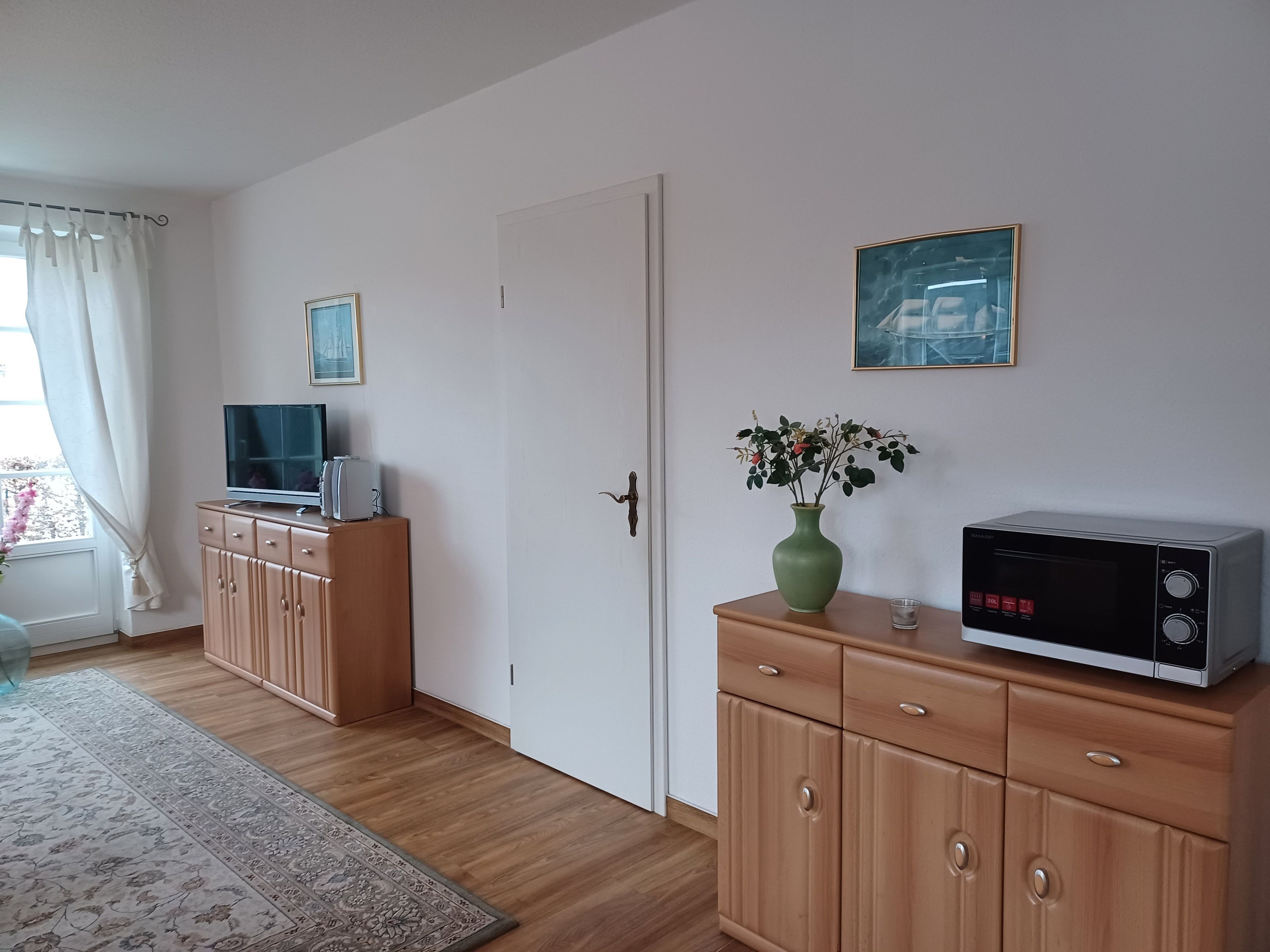 A room with a wooden cabinet, a microwave, a television, a vase with flowers, and a framed picture on the wall.