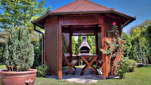 A wooden gazebo with a tiled roof, a built-in grill, and a seating area with two chairs. - Holiday apartment near the lake, with pool (Balatonmáriafürd����������������������������������������������������������������)