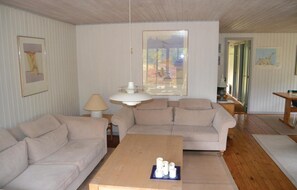 Un salon avec un canapé beige, une table basse en bois et une lampe. Les murs sont ornés de tableaux encadrés, et une fenêtre offre une vue sur les arbres.