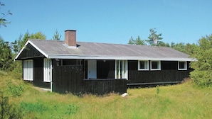 Ein einstöckiges, dunkel gefasstes Haus mit einem Satteldach, einem Schornstein und einer kleinen Veranda mit Fenstern in weißen Rahmen.