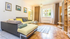 Ein modernes Wohnzimmer mit einer Couch, einem kleinen Tisch und einem Glastisch. An der Wand hängen drei gerahmte Kunstwerke, ein Fenster mit Vorhängen und ein Holzschrank.