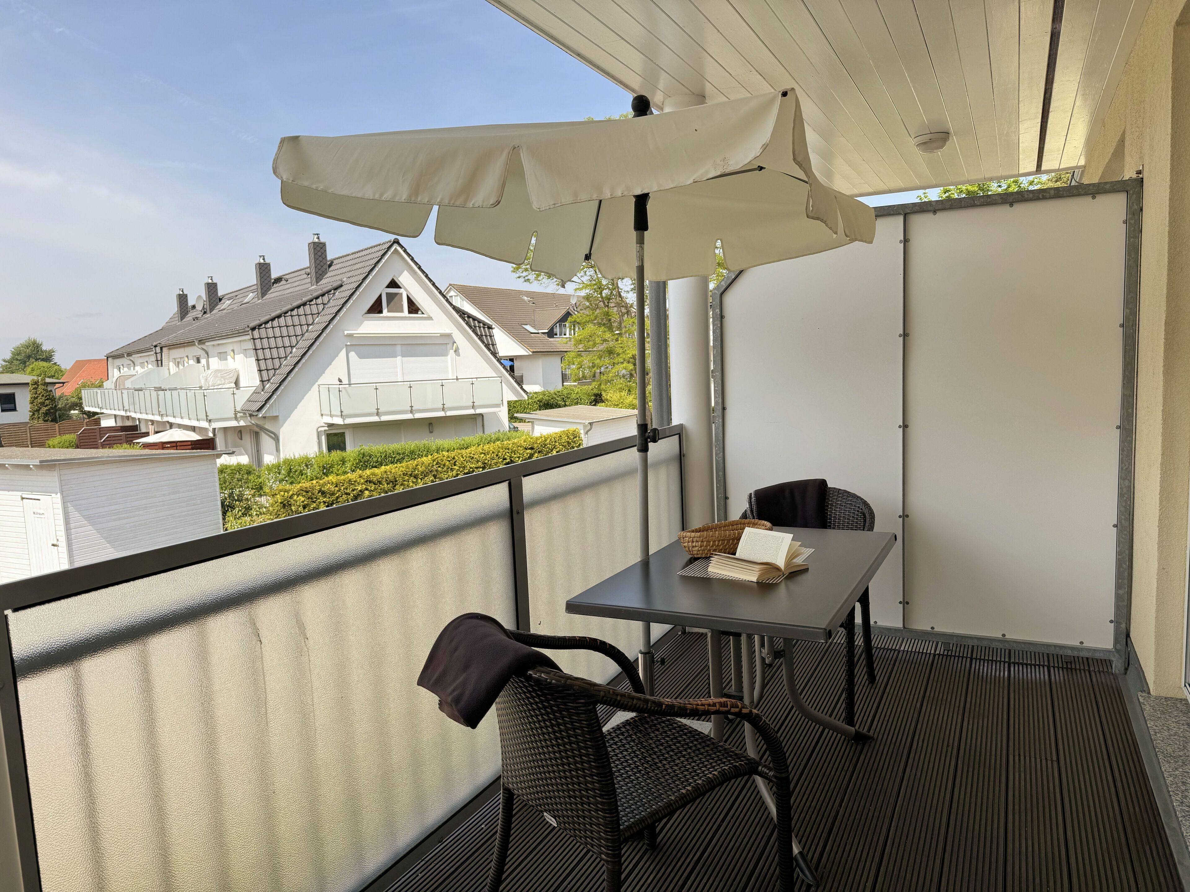 A balcony with a table and chairs, a white umbrella, and a view of houses in the background.