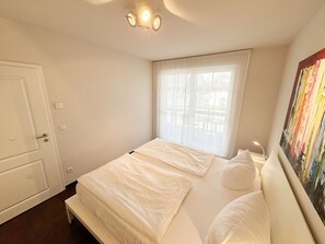 A bedroom with a bed, a window with sheer curtains, a door, and a painting on the wall. - F: Apartment Düne No. 09 - A jewel for your vacation! - Vacation apartment Düne (Thiessow)