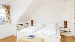 Quarto com teto inclinado, cama com roupa de cama branca, mesa de cabeceira com abajur e janela com cortinas.