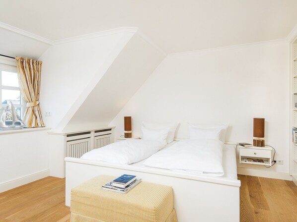 A bedroom with a sloped ceiling, a bed with white linens, a bedside table with a lamp, and a window with curtains.