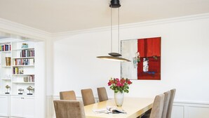 Uma área de refeições com uma mesa branca, seis cadeiras, um vaso com flores e uma estante cheia de livros e objetos decorativos.