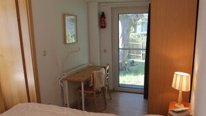 A bedroom with two beds, a wooden desk, a chair, a radiator, and a window with a view of the outside.