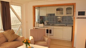 A compact living space with a sofa, armchair, and a small dining table with a bowl of fruit. A kitchen area with white cabinets and a microwave is visible in the background.