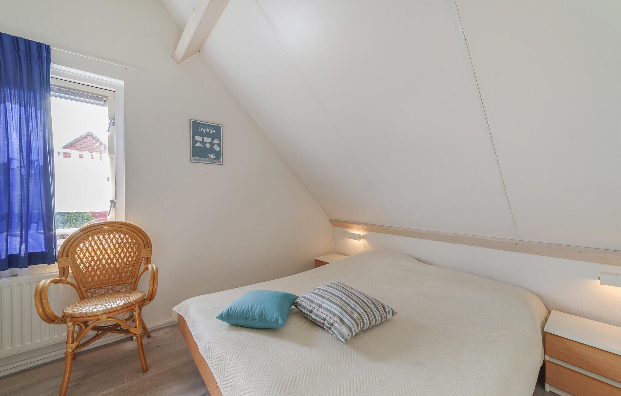 A bedroom with a sloped ceiling, a bed with two pillows, a wicker chair, and a window with blue curtains.