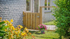 A wooden gate leading to a house with a brick wall, a window, and a potted plant with pink flowers.
