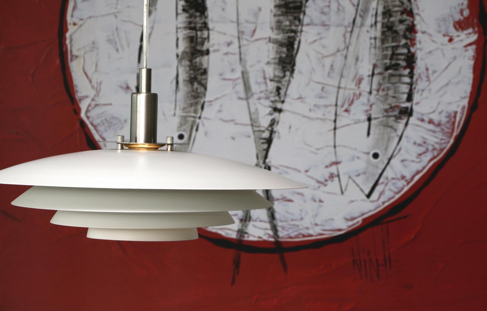A pendant light fixture with a white shade and a metallic base, mounted on a wall with a red background and a large abstract painting.