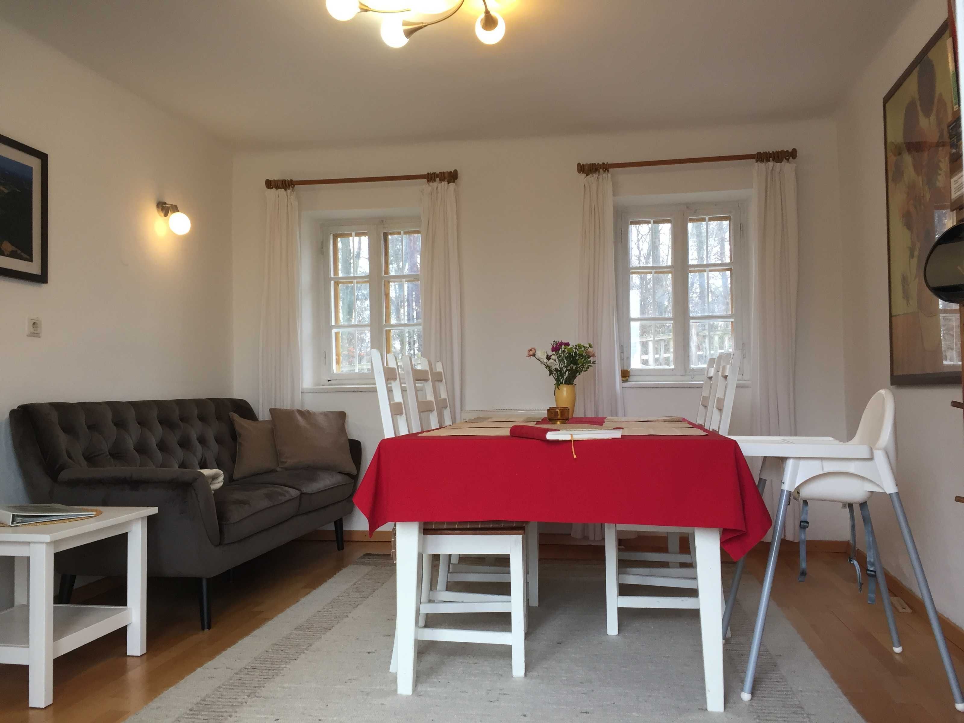 Ein Zimmer mit Esstisch für zwei Personen, einer Couch, einem kleinen Tisch mit einem Buch und zwei mit Vorhängen versehenen Fenstern.