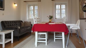 A room with a dining table set for two, a sofa, a small table with a book, and two windows with curtains.