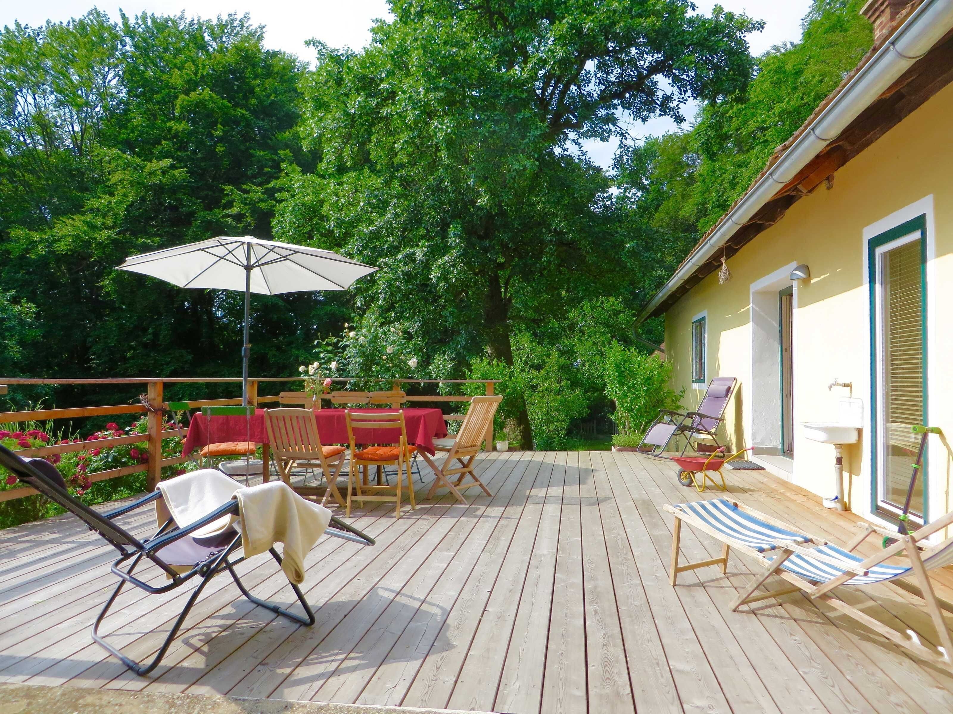 A wooden deck with outdoor furniture, including chairs and a table, surrounded by lush greenery.
