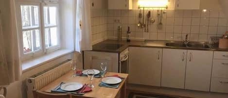 Un coin repas avec une table dressée pour deux personnes, une fenêtre avec des rideaux et une cuisine dotée de meubles blancs et d’un plan de travail.