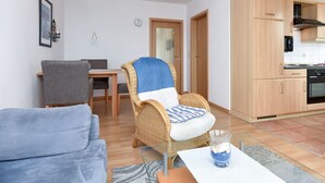 Un salon comprenant un canapé, un fauteuil en osier, une table à manger et un coin cuisine avec des meubles et des appareils électroménagers en bois.