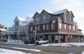 A multi-story building with a flat roof, a visible entrance, and a parking area with cars. - Lee - house harbor view (Esens)