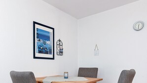 Un coin repas avec une table en bois, deux chaises grises à revêtement rembourré, un tableau encadré, une horloge murale et un petit luminaire fixé au mur.