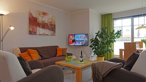 Ein Wohnzimmer mit einer Couch, Sesseln, einem Couchtisch, einem Fernseher, einer Pflanze und einem Fenster mit Vorhängen.