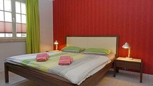 Ein ordentlich gemachtes Bett mit Kissen und Handtüchern, ein Nachttisch mit Lampe und ein Fenster mit Jalousien.