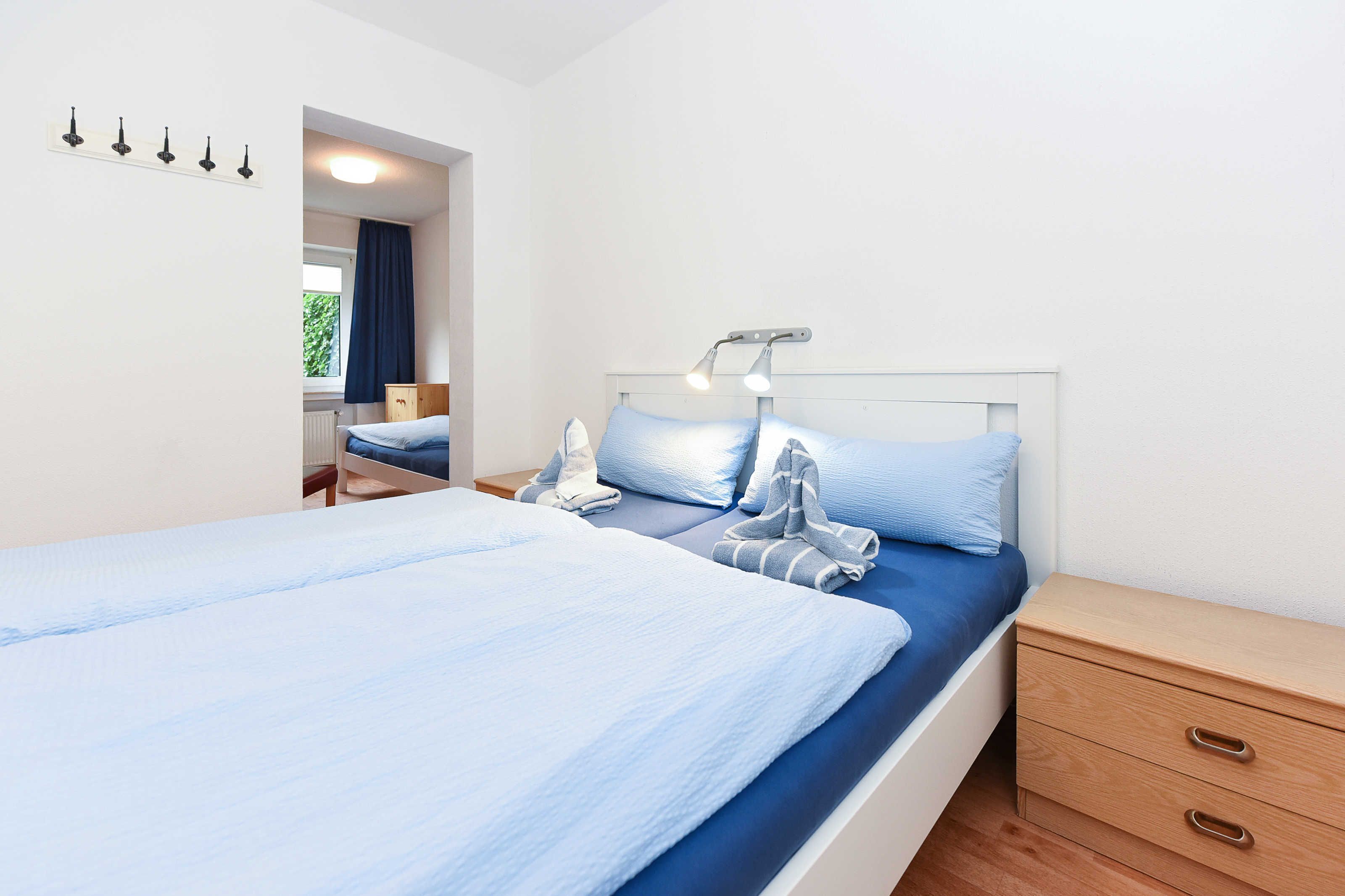 Ein ordentlich bezogenes Bett mit blauer Matratze und Kissen, eine Nachttischlampe, ein Nachttisch aus Holz mit Schubladen und ein Fenster mit Vorhängen.