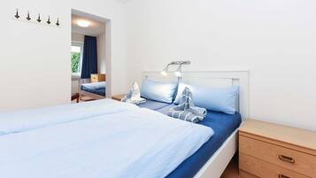 Ein ordentlich bezogenes Bett mit blauer Matratze und Kissen, eine Nachttischlampe, ein Nachttisch aus Holz mit Schubladen und ein Fenster mit Vorhängen.