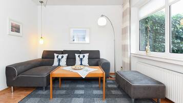 Ein modernes Wohnzimmer mit einem grauen Ecksofa, einem Holztisch und einer grauen Bank. Ein großes Fenster bietet Blick auf Grünflächen.