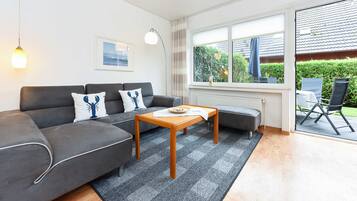 Ein modernes Wohnzimmer mit einem grauen Sofa, einem Holztisch und Blick auf einen Garten durch große Fenster.