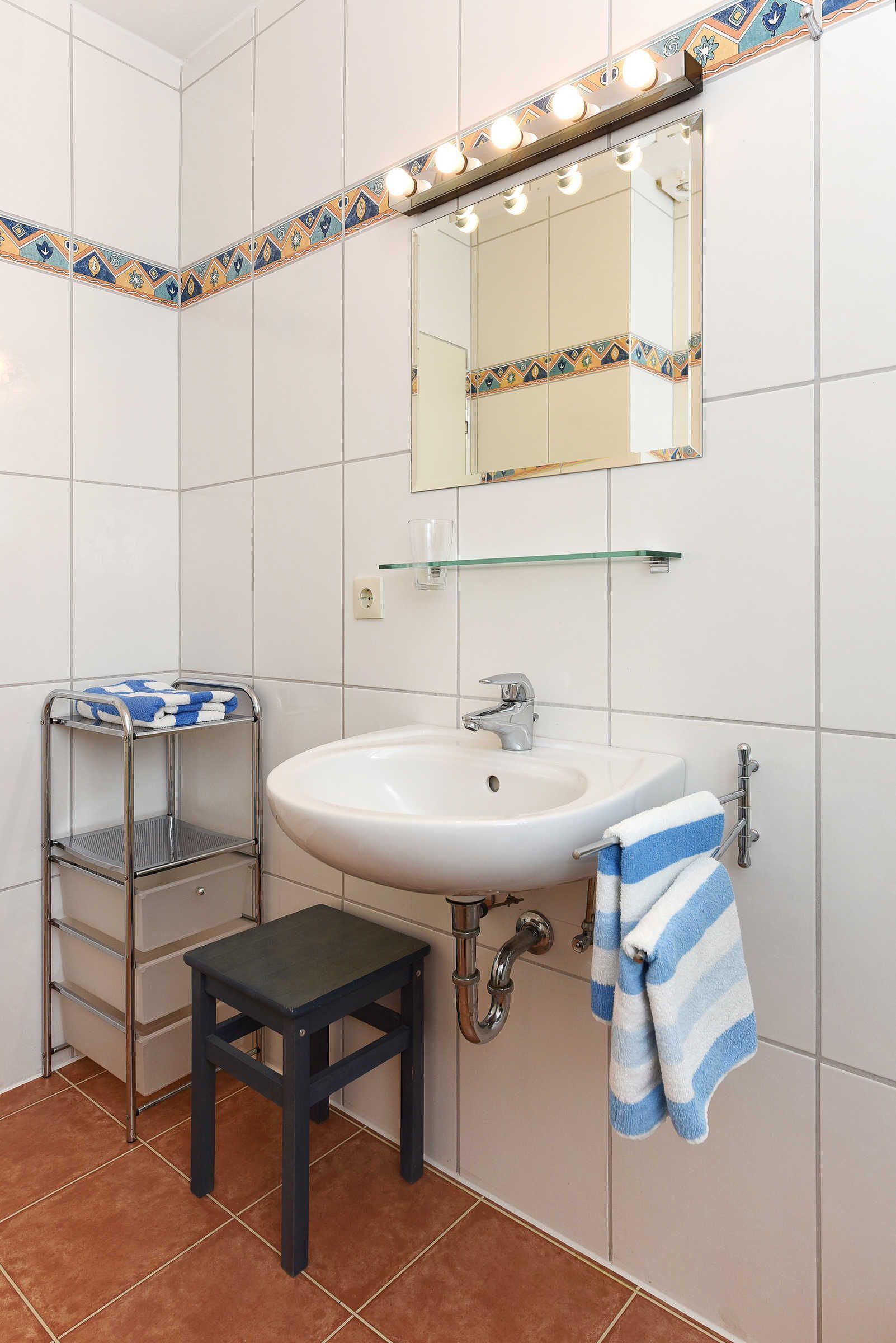 A bathroom with a white sink, a mirror with lights, a towel rack, and a shelf with towels.