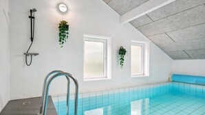 Una piscina interior moderna con baldosas azules cristalinas, una escalera metálica y dos ventanas con plantas colgantes.