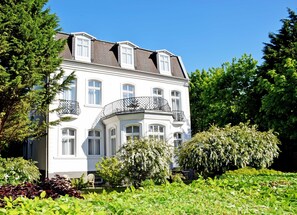 A white building with multiple windows and balconies, surrounded by lush greenery and trees. - 03 Multi-bed room (2-room) - Villa in the Ostseebad Baabe (Baabe)