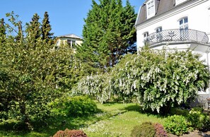 A lush garden with a variety of trees and shrubs, a white building with a balcony, and a clear blue sky.