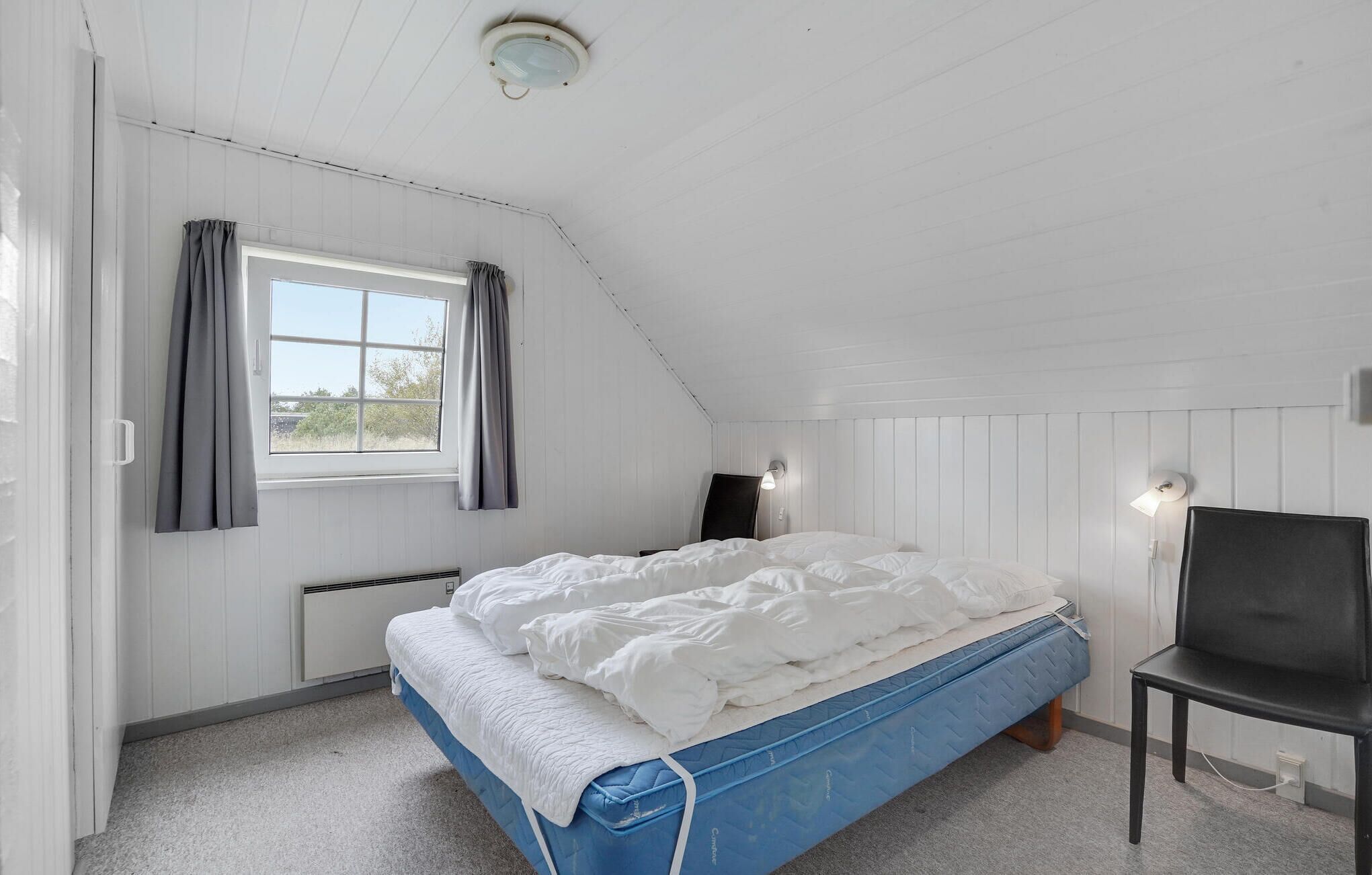 Een slaapkamer met een schuine kap, een bed met wit beddengoed, een blauwe matras, een stoel, een televisie en een raam met gordijnen.