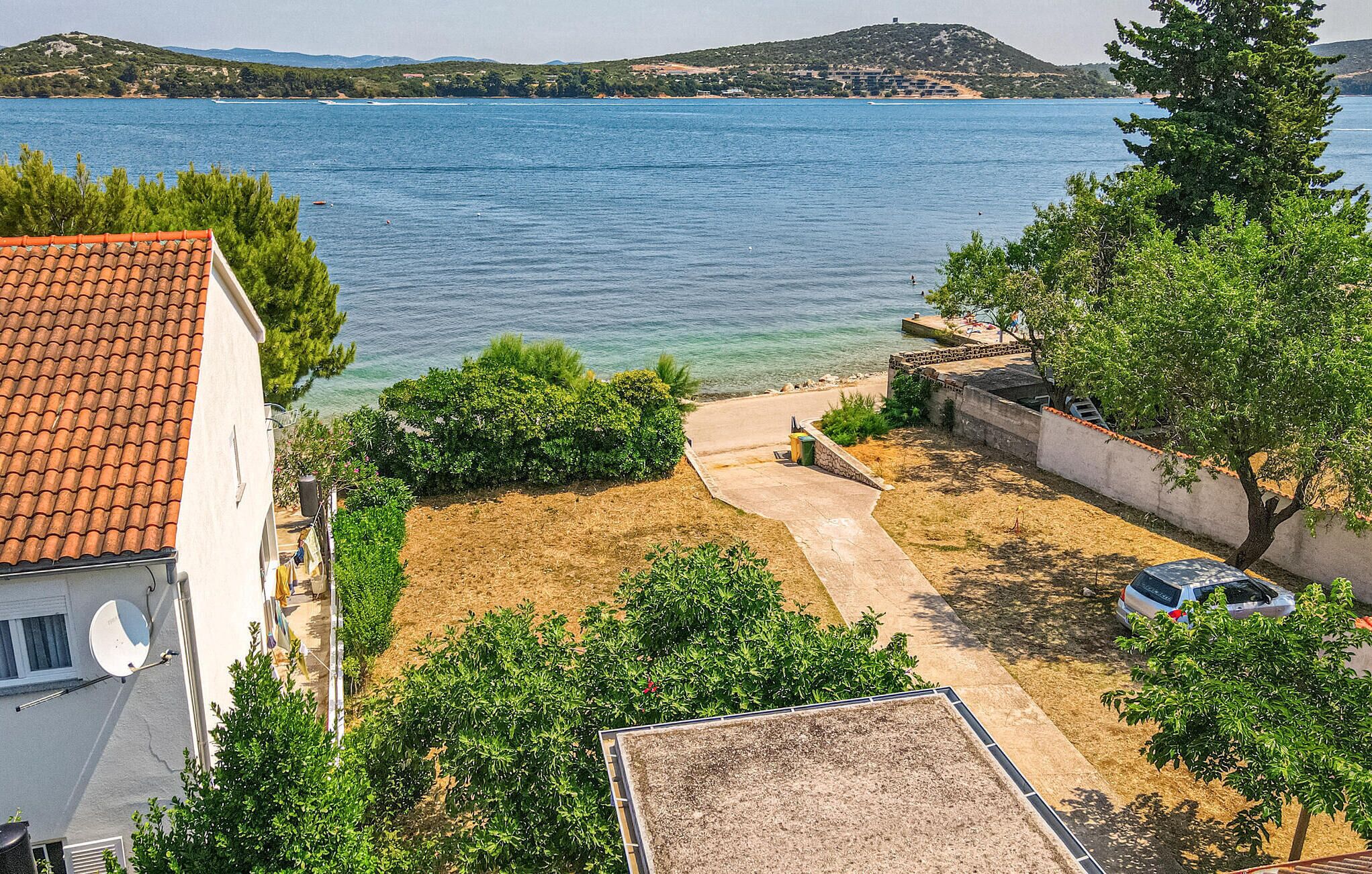 A coastal property with a paved driveway leading to a sandy area, surrounded by greenery and a view of the sea.